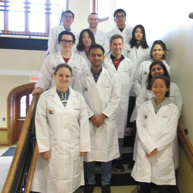 12 students in white lab coats pose in stairwell