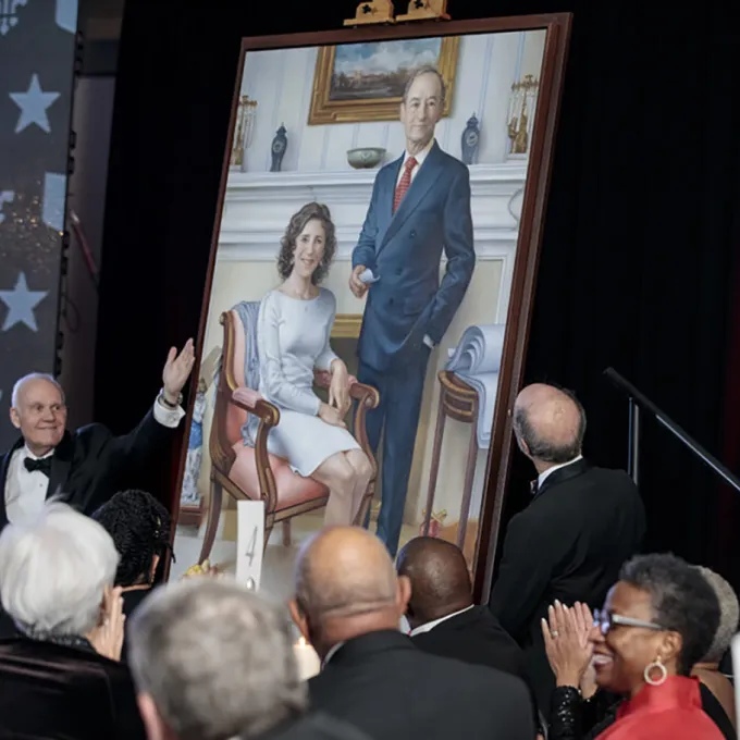 a man in tuxedo gestures to a large portrait of the Wrightons